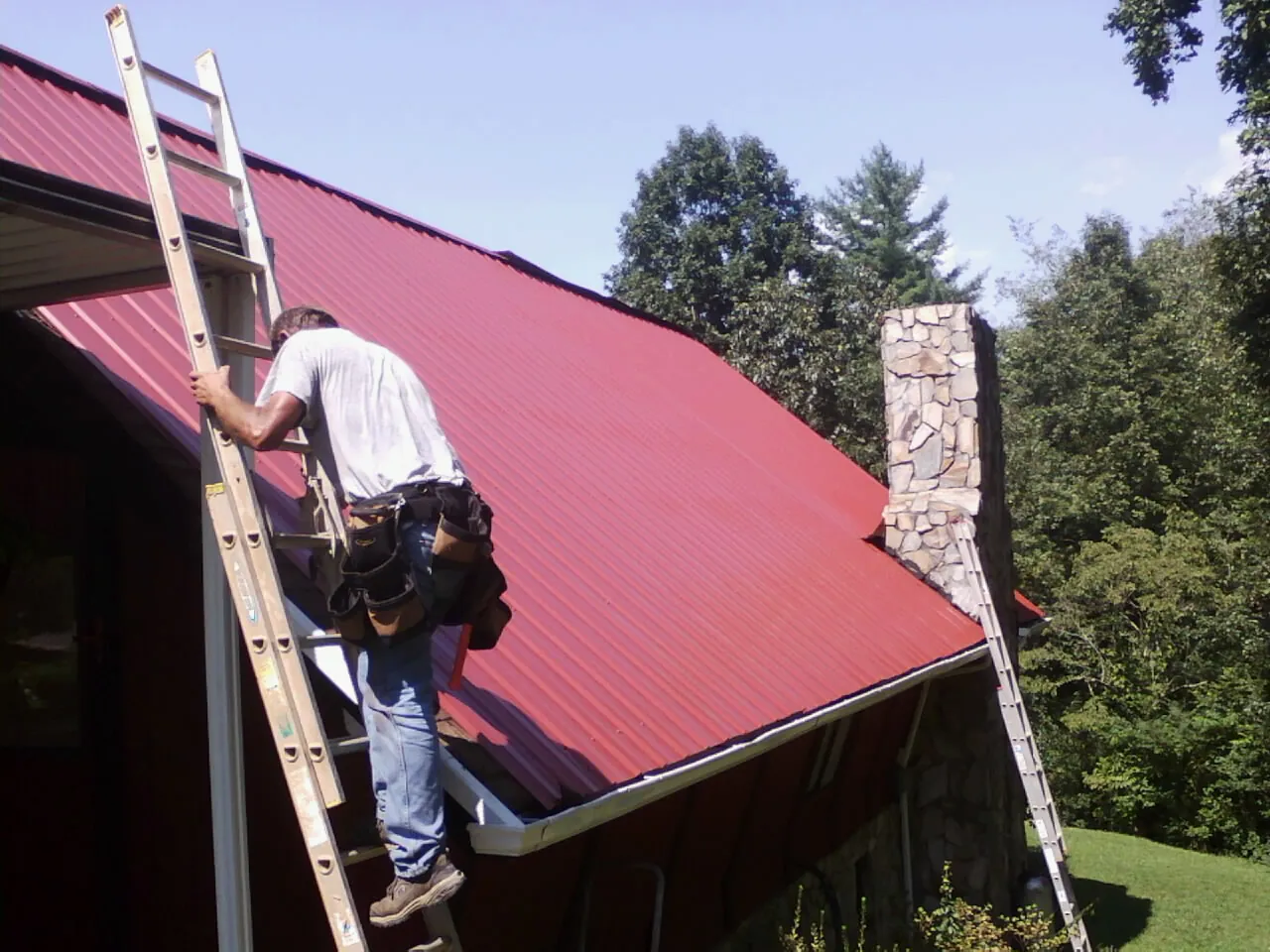 Roofer on a ladder beside a red metal roof and stone chimney — Soffit & Fascia Repair by Roofing Brothers in Redding